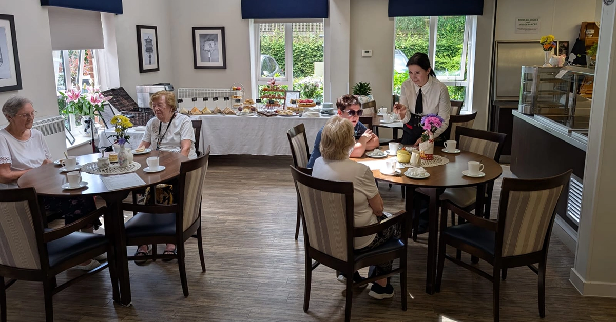 Four elderly women sit at tables in a bright café, with tea, flowers, and pastries; a woman serves drinks.