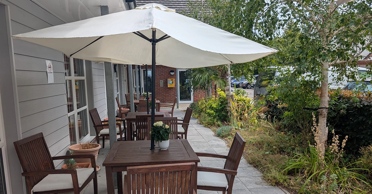 A patio with wooden tables, chairs, and a large white parasol next to a garden and building walkway.